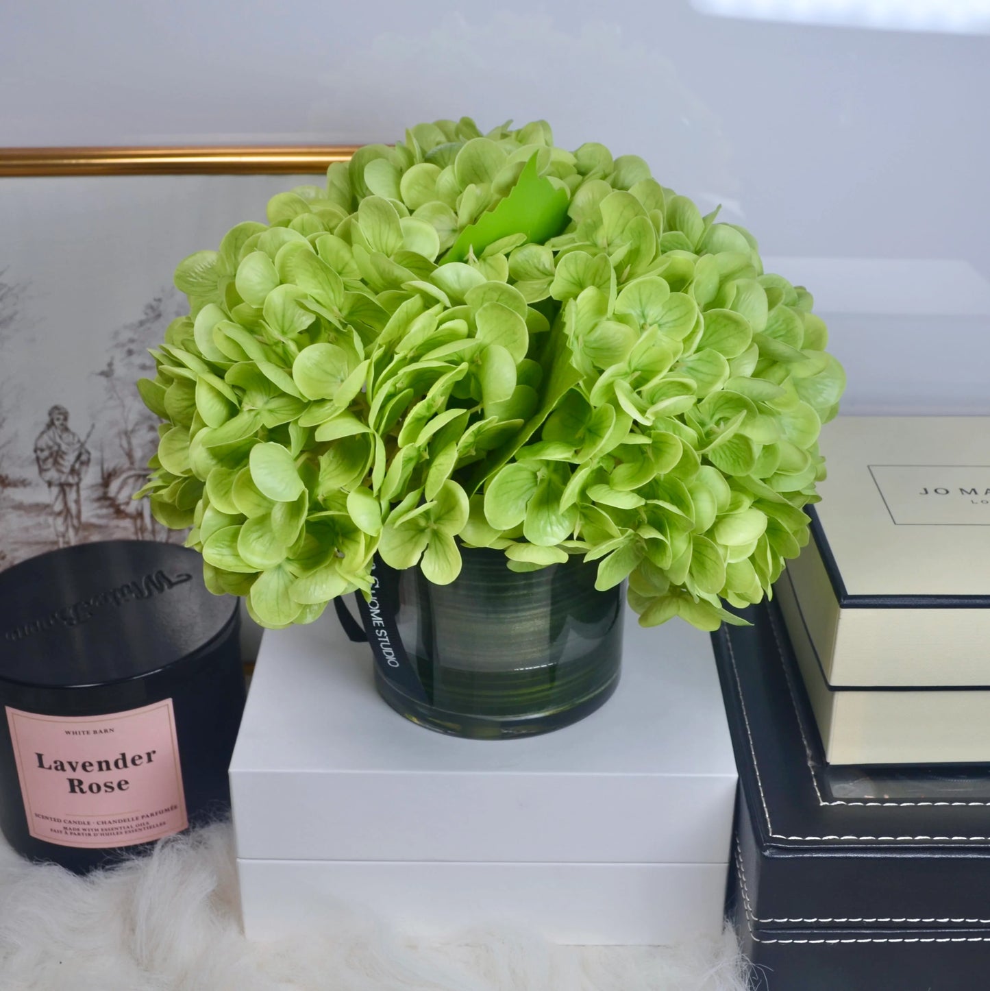 Green hydrangea flower arrangement on a white surface with books and a candle in the background.