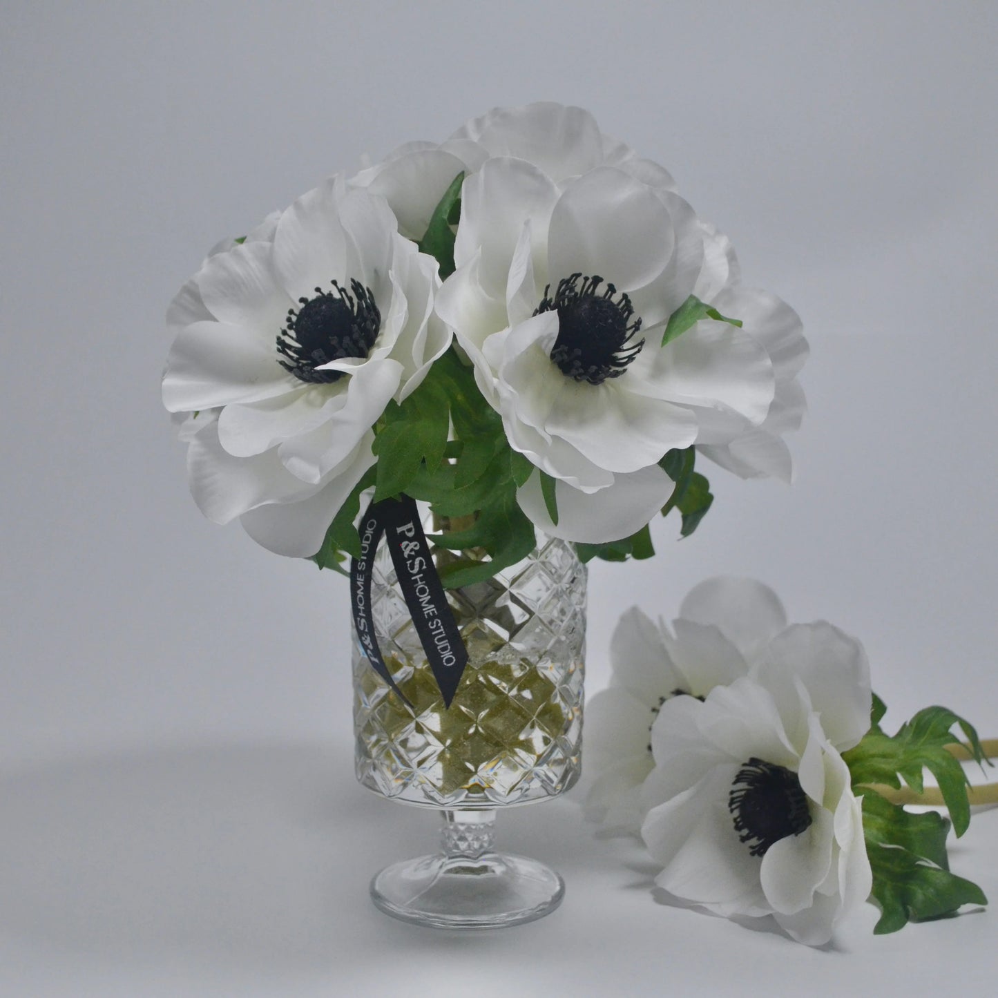 White flowers with green leaves in a clear glass vase on a light gray background