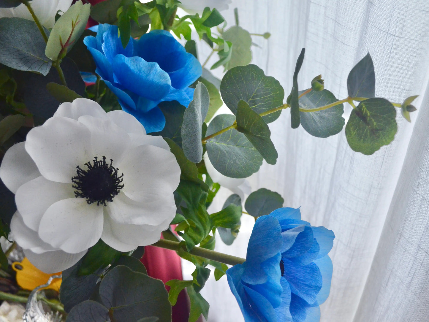 Bouquet of white anemones and blue flowers with green leaves against a white curtain background.