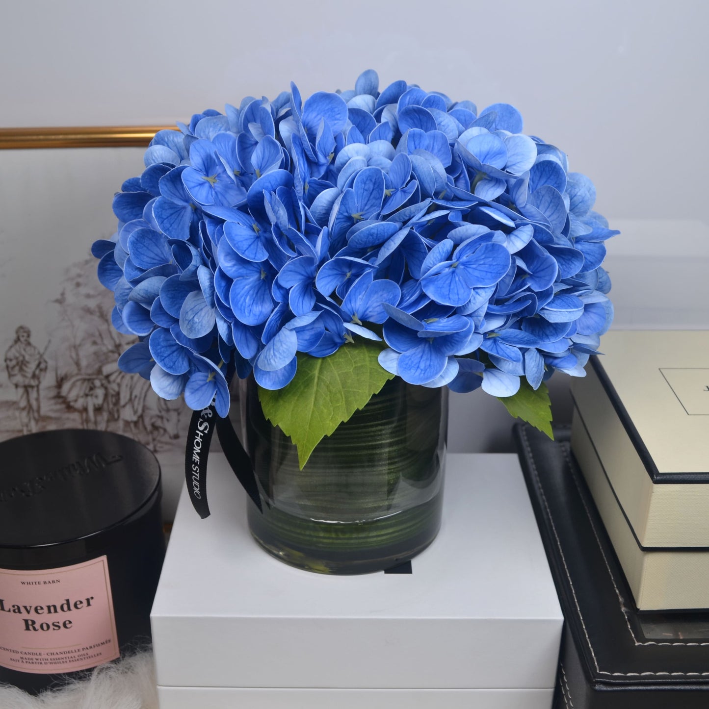 Bouquet of blue hydrangeas in a clear vase on a white surface with books and a candle in the background.