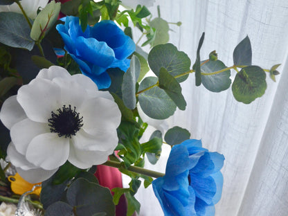 Bouquet of white anemones and blue flowers with green leaves against a white curtain background.